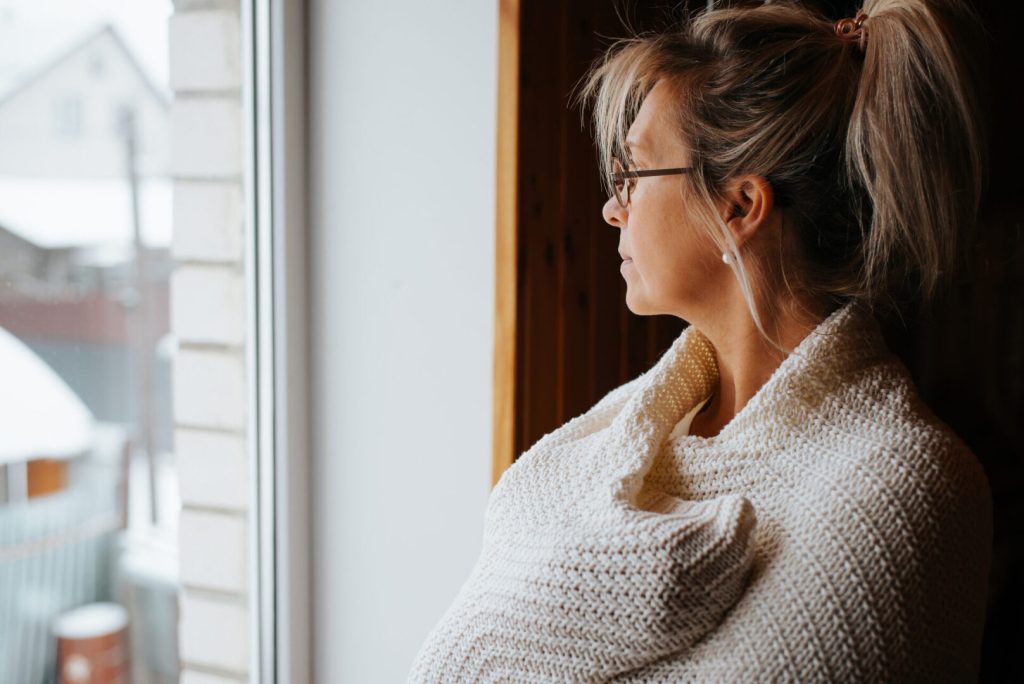 Side view portrait of sad lonely adult caucasian woman wrapped in warm cozy knitted blanket standing by window and looking away, indoors. Winter blues.